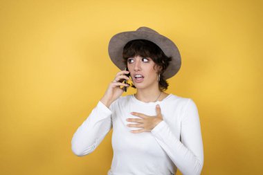 Sarı arka planda şapka takan beyaz bir kadın telefonda endişeli bir ifadeyle konuşuyor.