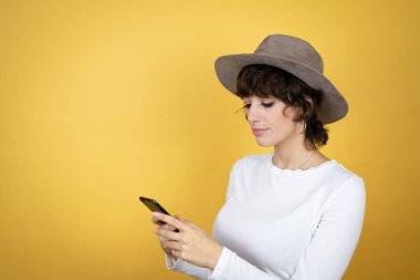 İzole edilmiş sarı arka planda şapka takan beyaz bir kadın telefonuyla mesajlaşıyor.