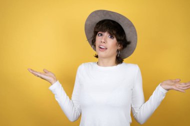 İzole edilmiş sarı arka planda şapka takan beyaz bir kadın. Kolları ve elleri havada, kafası karışmış bir ifade.