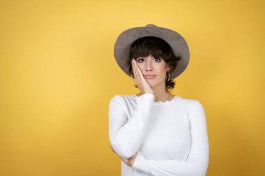 Sarı arka planda şapka takan genç beyaz kadın çapraz kollardan sıkılmış ve yorgun görünüyor.