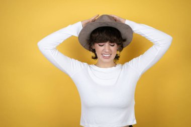 Kafkasyalı genç bir kadın, sarı arka planda izole edilmiş bir şapka takıyor. Baş ağrısı çekiyor. Çaresiz ve stresli.