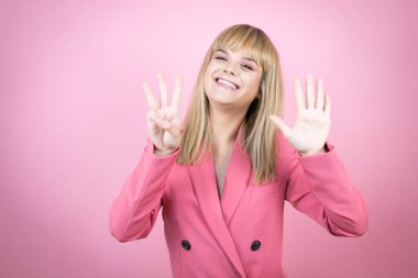 İzole edilmiş pembe arka planda genç beyaz bir kadın sekiz numaralı parmaklarıyla gülümseyerek kendinden emin ve mutlu bir şekilde onu işaret ediyor.