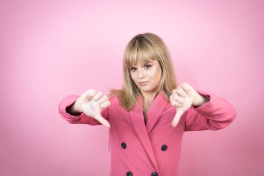 Kafkasyalı genç bir kadın, pembe arka planda, kızgın yüzlü, negatif işaretli, baş parmakları aşağıda.