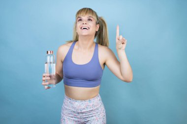 Spor kıyafetleri giymiş genç bir kadın mavi arka planda bir şişe su tutuyor. Şaşırmış ve şaşırmış bir şekilde yukarı bakıyor ve parmaklarıyla işaret ediyor ve kollarını kaldırıyor.