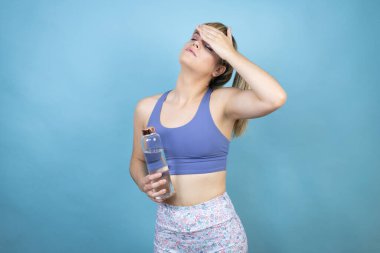 Spor kıyafetleri giyen genç bir kadın izole edilmiş mavi arka planda bir şişe su tutuyor. Hastalık, ateş, grip ve soğuk algınlığı için alnına dokunuyor.