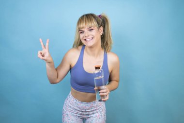 Spor kıyafetleri giymiş genç bir kadın izole edilmiş mavi arka planda bir şişe su tutuyor ve iki numarayı gösterirken kendinden emin ve mutlu bir şekilde gülümsüyor.