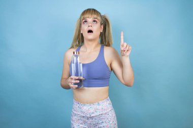 Spor kıyafetleri giymiş genç bir kadın mavi arka planda bir şişe su tutuyor. Şaşırmış ve şaşırmış bir şekilde yukarı bakıyor ve parmaklarıyla işaret ediyor ve kollarını kaldırıyor.