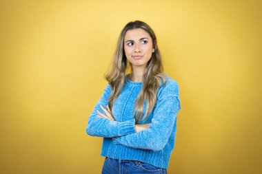 Sarı arka planda duran uzun saçlı güzel sarışın kadın çapraz kollardan sıkılmış ve yorgun göründüğünü düşünüyor.