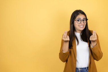 İzole edilmiş sarı arka planda ceket giyen genç ve güzel bir kadın. Elleriyle para hareketi yapıyor. Maaş ödemesi istiyor.