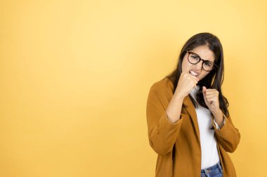 Sarı arka planda ceket giyen genç ve güzel bir kadın dövüşmek için yumruk, agresif ve öfkeli saldırı, tehdit ve şiddet.