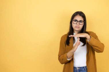İzole edilmiş sarı arka planda ceketini giyen genç ve güzel bir kadın. Elleriyle jest yapıyor, sinirli ve ciddi bir yüz ifadesi takınıyor.