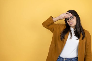 İzole edilmiş sarı arka planda ceket giyen genç ve güzel bir kadın. Hastalık ve ateş için alnına dokunuyor. Grip ve soğuk algınlığı için.