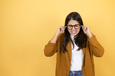 İzole edilmiş sarı arka planda ceket giyen genç ve güzel bir kadın yüksek sesli müzik sesi için kulakları parmaklarıyla kapatıyor. Sağır kavramı.