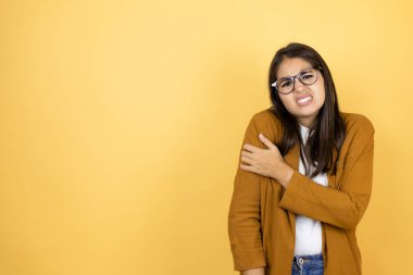 İzole edilmiş sarı arka planda ceket giyen genç ve güzel bir kadın. Omzunda ağrı ve acı dolu bir ifade var.