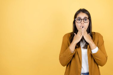 İzole edilmiş sarı arka planda ceket giyen genç ve güzel bir kadın. Elleri ağzında ve şaşırmış.