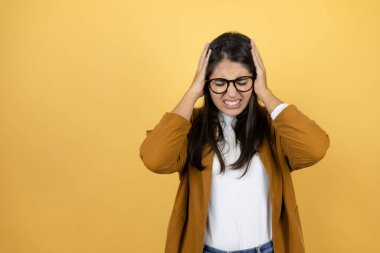 Sarı arka planda ceket giyen genç ve güzel bir kadın baş ağrısı ve stresten muzdarip çünkü elleri başının üstünde acı ve migren var.