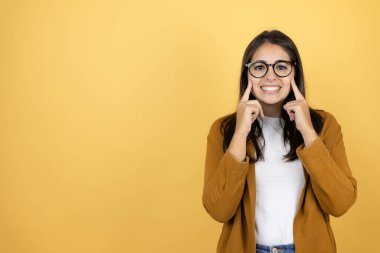 İzole edilmiş sarı arka planda ceket giyen genç ve güzel bir kadın gülümsüyor ve parmaklarıyla ve ağzıyla işaret ediyor.