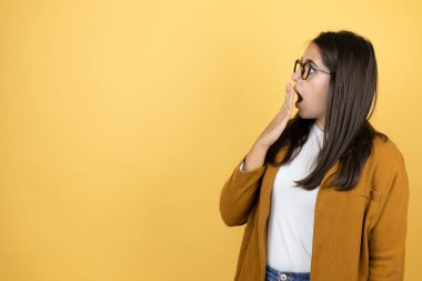 İzole edilmiş sarı arka planda ceket giyen genç ve güzel bir kadın. Elleri ağzında ve şaşırmış, yan bakıyor.