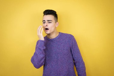 İzole edilmiş sarı arka plandaki genç beyaz adam, ağzını eliyle kapatmaktan sıkılmış. Huzursuz ve uykulu..
