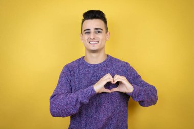 İzole edilmiş sarı arka planda genç beyaz bir adam. Sevgiyle gülümsüyor. Elleriyle kalp sembolü ve şekli gösteriyor.