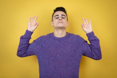 Kafkasyalı genç adam izole edilmiş sarı arka planda rahatla ve gözleri kapalı gülümse parmaklarıyla meditasyon hareketi yapıyor.