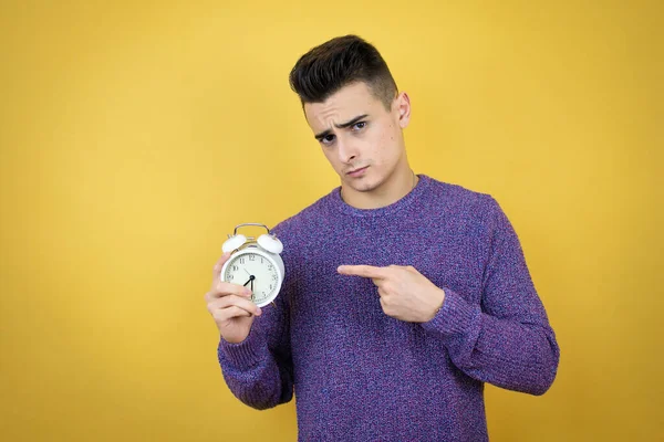 Young Caucasian Man Isolated Yellow Background Serious Holding Pointing Clock — Stock Photo, Image