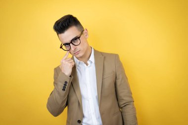İzole edilmiş sarı arka planda genç bir iş adamı. Gözünü dikip jestini izliyor. Şüpheli bir ifade.