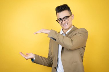 İzole edilmiş sarı arka planda, büyük ve büyük boyut işareti gösteren el işaretleri olan genç bir iş adamı. Gülümsüyor. Ölçüm konsepti.