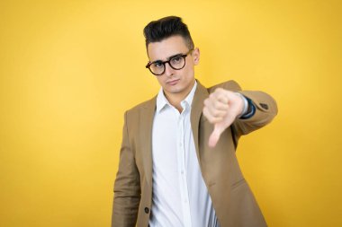 Sinirli yüzüyle izole edilmiş sarı arka planda genç bir iş adamı. Negatif işaretler baş parmağın aşağıda olduğunu gösteriyor.