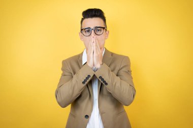 Genç bir iş adamı, sarı arka planda, elleri ağzında ve şaşırmış.