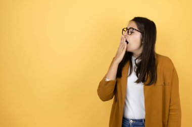 İzole edilmiş sarı arka plan üzerine ceket giyen genç ve güzel bir kadın ağzıyla ağzını kapatmaktan sıkılmış. Huzursuz ve uykulu..