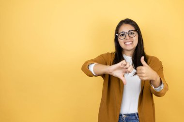 İzole edilmiş sarı arka planda ceket giyen genç ve güzel bir kadın baş parmak hareketi yapıyor, anlaşmazlık ve anlaşma ifadesi. Çılgın çatışma