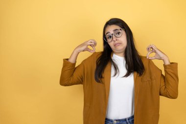 İzole edilmiş sarı arka plan üzerine ceket giyen genç ve güzel bir kadın tişörtünü başarılı bir ifadeyle tutuyor.