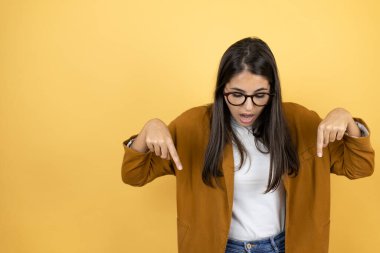 İzole edilmiş sarı arka plan üzerine ceket giyen genç ve güzel bir kadın şaşırmış, aşağı bakıyor ve parmaklarıyla işaret ediyor ve kollarını kaldırıyor.
