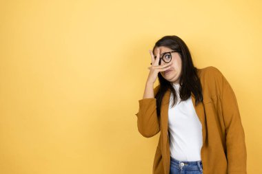 İzole edilmiş sarı arka planda ceket giyen genç ve güzel bir kadın. Şoka girmiş, yüzünü ve gözlerini kapamış, utangaç ifadesiyle parmaklarından bakıyor.