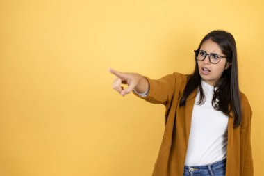 İzole edilmiş sarı arkaplan üzerine ceket giyen genç ve güzel bir kadın parmağıyla işaret ederek ileriyi işaret ediyor, ağzı açık hayret verici bir ifade, ön tarafta bir şey.