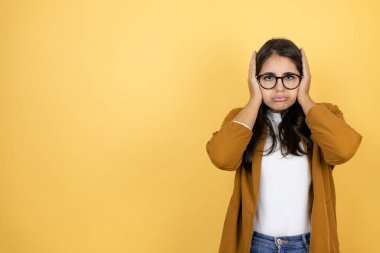 İzole edilmiş sarı arka planda ceket giyen genç ve güzel bir kadın yorgun ve elleri başının üstünde sıkılmış olarak düşünüyor.