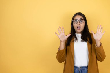 İzole edilmiş sarı arka plan üzerinde ceketini giyen genç ve güzel bir kadın korku ifadesinden korkuyor ve dehşete düşüyor. El hareketlerini durdur, şok içinde bağır. Panik konsepti.