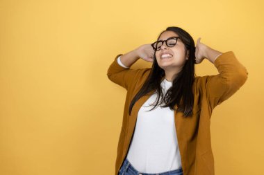 İzole edilmiş sarı arka planda ceket giyen güzel bir kadın rahatlıyor ve esniyor, kolları ve elleri başının arkasında ve boynunun arkasında mutlu bir şekilde gülümsüyor.