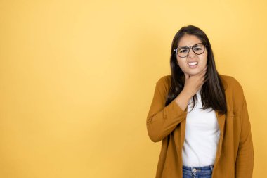 İzole edilmiş sarı arka planda ceket giyen genç ve güzel bir kadın ağrıyan boynuna dokunuyor, grip, mantar ve enfeksiyon yüzünden boğazı ağrıyor.