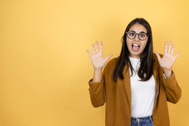 İzole edilmiş sarı arka planda ceket giyen genç ve güzel bir kadın on numaralı parmaklarıyla işaret ederken kendinden emin ve mutlu bir şekilde gülümsüyor.