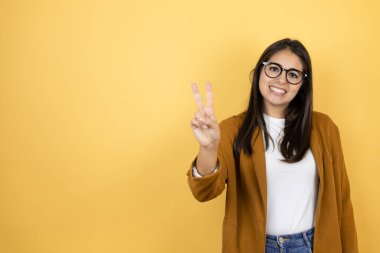 İzole edilmiş sarı arka planda ceket giyen genç ve güzel bir kadın iki numaralı parmaklarıyla gülümseyerek kendinden emin ve mutlu bir şekilde onu işaret ediyor.