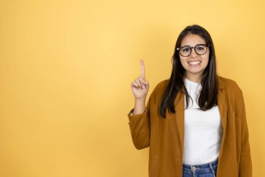 İzole edilmiş sarı arka planda ceket giyen genç ve güzel bir kadın bir numaralı parmaklarıyla gülümseyerek kendinden emin ve mutlu bir şekilde onu işaret ediyor.