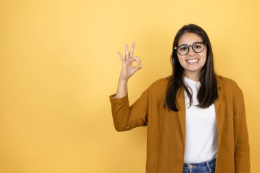 İzole edilmiş sarı arka plan üzerine ceket giyen genç ve güzel bir kadın tamam işareti yapıyor parmaklarıyla ve gülümseyerek, mükemmel bir sembol.
