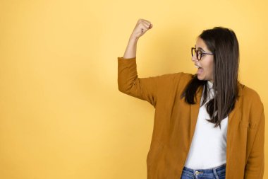 İzole edilmiş sarı arka planda ceket giyen güzel bir kadın gururla gülümseyen kol kaslarını gösteriyor.
