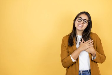 İzole edilmiş sarı arka planda ceket giyen genç ve güzel bir kadın elleri göğsünde ve yüzünde minnet dolu bir jest ile gülümsüyor..