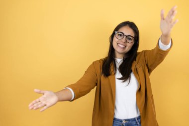 İzole edilmiş sarı arka planda ceket giyen genç ve güzel bir kadın kameraya sarılıp gülümseyerek bakıyor.