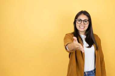 İzole edilmiş sarı arka planda ceket giyen genç ve güzel bir kadın gülümseyerek selamlama ve hoş geldin demek için tokalaşıyor.