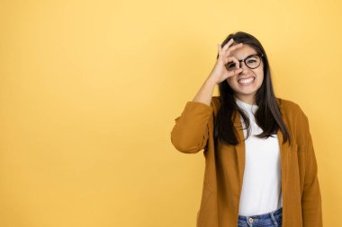 İzole edilmiş sarı arka planda ceketini giyen genç ve güzel bir kadın gülümseyen yüzüyle, parmaklarından bakan gözleriyle şok olmuş bir jest yapıyor.