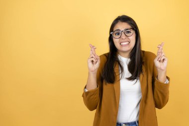 İzole edilmiş sarı arka plan üzerinde ceket giyen genç ve güzel bir kadın umut dolu bir gülümsemeyle işaret ediyor ve bakıyor.
